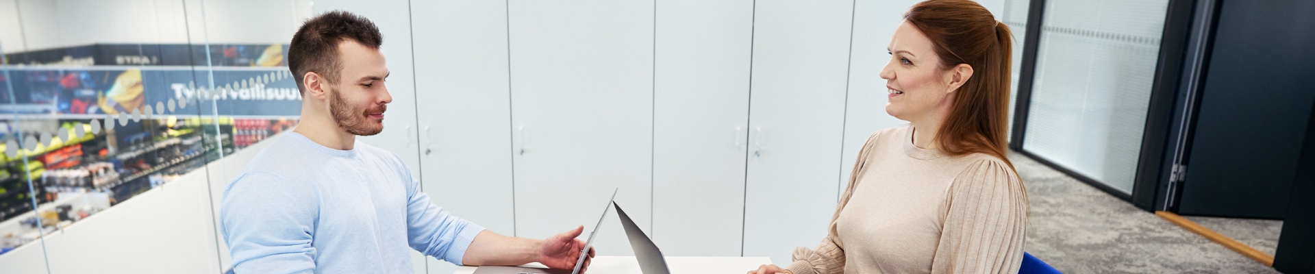 A man and a woman are sitting across from each other in an office at a conference table, each with a laptop in front of them. They are smiling while having a conversation.