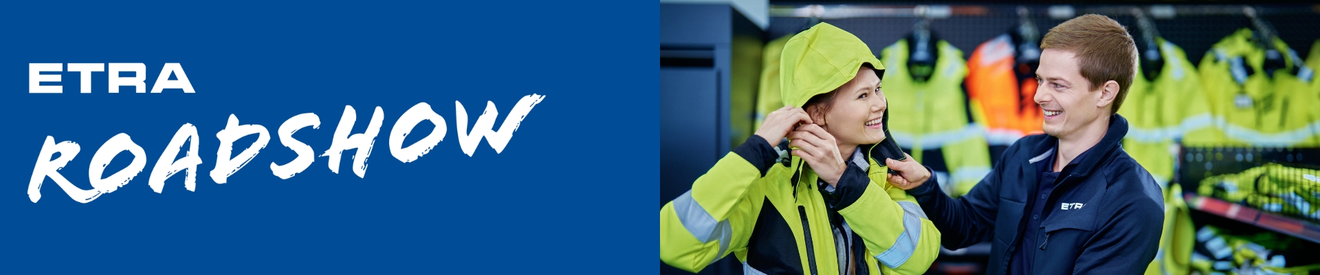 Kaksi hymyilevää henkilöä sovittamassa työturvallisuusvaatteita ETRA Roadshow -tapahtumassa, taustalla näkyy värikkäitä työvaatteita.