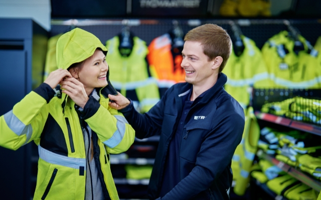An Etra staff member helps a customer try on a high-visibility work jacket.