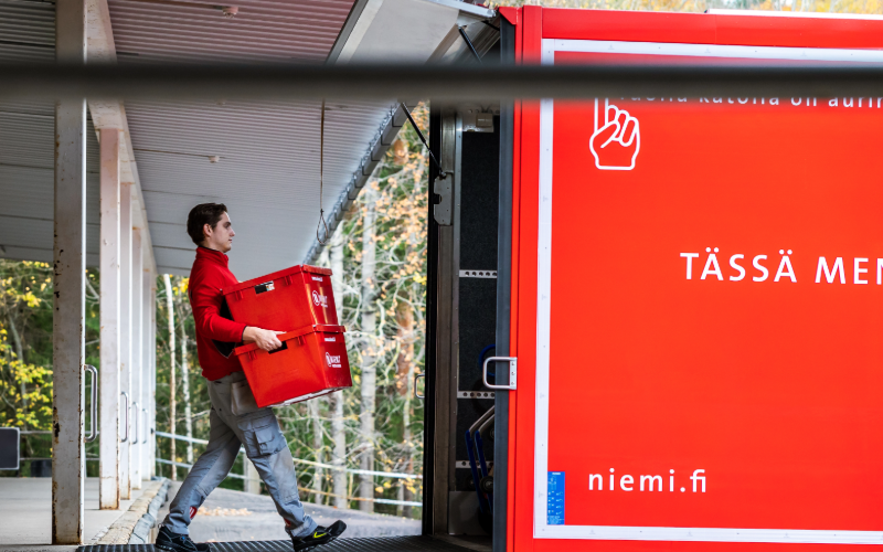 Niemi Palveluiden muuttotyöntekijä kantaa kahta punaista muuttolaatikkoa kuorma-auton takalaitanostinta pitkin auton sisään lastauslaiturin luona.