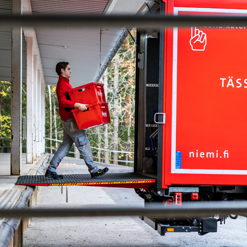 Niemi Palveluiden muuttotyöntekijä kantaa kahta punaista muuttolaatikkoa kuorma-auton takalaitanostinta pitkin auton sisään lastauslaiturin luona.