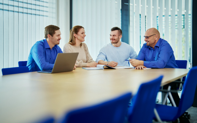Four people are sitting around a modern conference table, engaged in conversation and smiling. Two of them are wearing blue dress shirts, one is wearing a beige sweater, and one is wearing a light blue sweater. There is a laptop on the table.