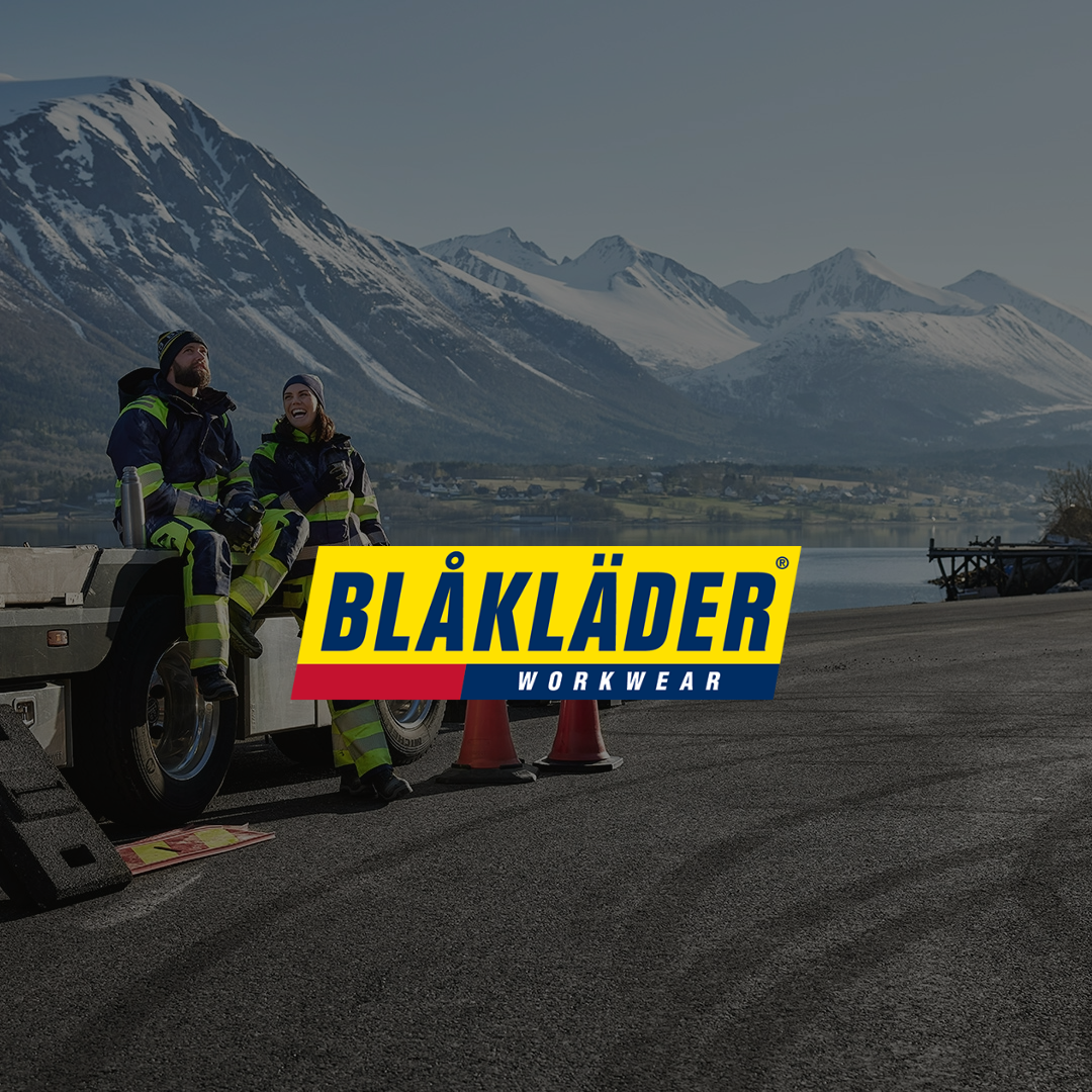 Kaksi henkilöä istuu työvaatteissa raskaan ajoneuvon päällä asfalttitiellä, taustalla näkyy vuoristomaisema ja järvi. Kuvan keskellä on BLÅKLÄDER WORKWEAR -logo.