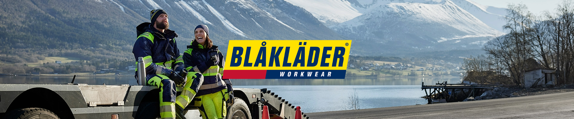 Kaksi henkilöä yllään Blåkläderin työvaatteet istuu perävaunun reunalla talvisessa vuoristomaisemassa järven rannalla. Kuvan keskellä on Blåkläder Workwear -logo keltaisella, sinisellä ja punaisella taustalla.