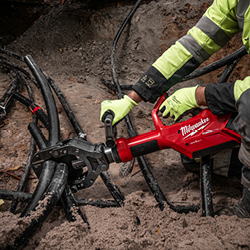 Rakennustyöntekijä käyttää Milwaukee-merkkistä hydraulista leikkuria leikatakseen maassa olevia paksuja sähkö- tai tietoliikennekaapeleita. Työntekijällä on neonkeltaiset heijastavat työtakit ja käsineet.