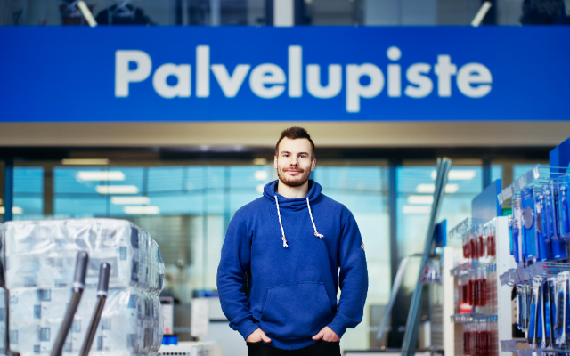Nuori mies seisoo myymälän palvelupisteen edessä sinisessä hupparissa. Taustalla näkyy myymälähyllyjä ja tuotteita, korostaen asiakaspalvelun saavutettavuutta ja nykyaikaista myymäläympäristöä.