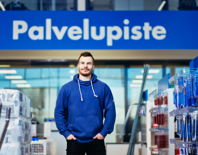 Nuori mies seisoo myymälän palvelupisteen edessä sinisessä hupparissa. Taustalla näkyy myymälähyllyjä ja tuotteita, korostaen asiakaspalvelun saavutettavuutta ja nykyaikaista myymäläympäristöä.