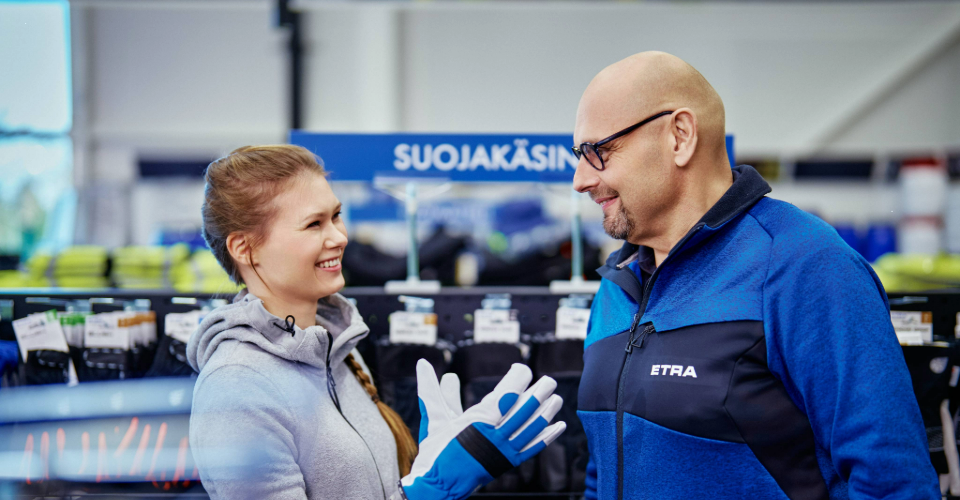 A smiling woman tries on work gloves and talks with store staff at Etra's protective gloves section.