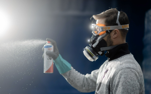 A man wearing protective gear uses a spray can and respirator in a work environment.