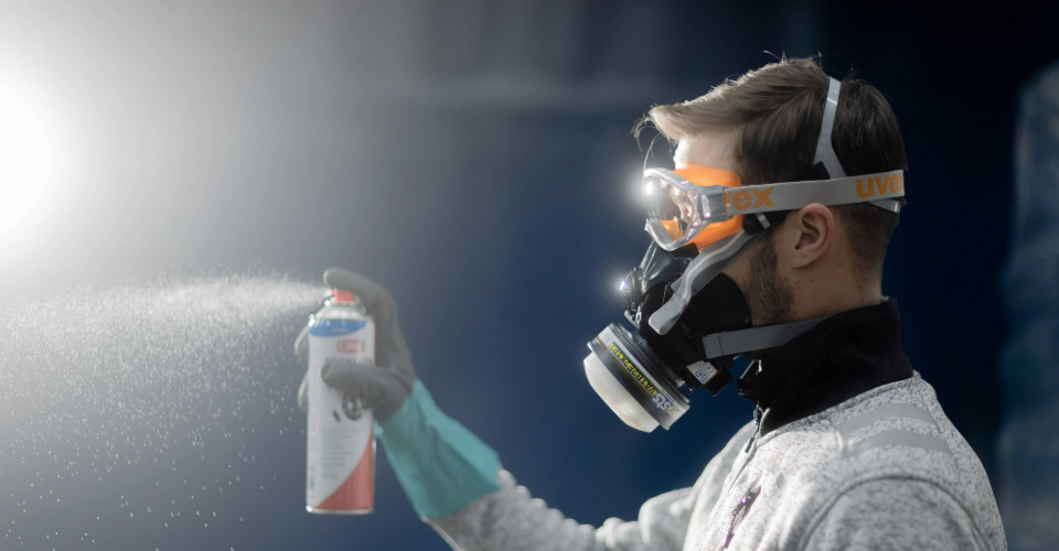  A man wearing protective gear uses a spray can and respirator in a work environment.