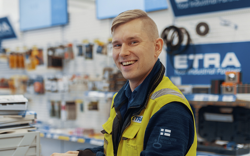 Hymyilevä mies asiakaspalvelija seisoo myymälässä ETRA-kyltin edessä, päällään keltainen huomioliivi ja sininen työasu, taustalla hyllyjä täynnä työkaluja ja tarvikkeita.