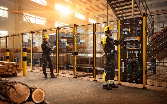 Wood industry production facility where workers in safety gear operate near a protected machine line and use wall-mounted tools.