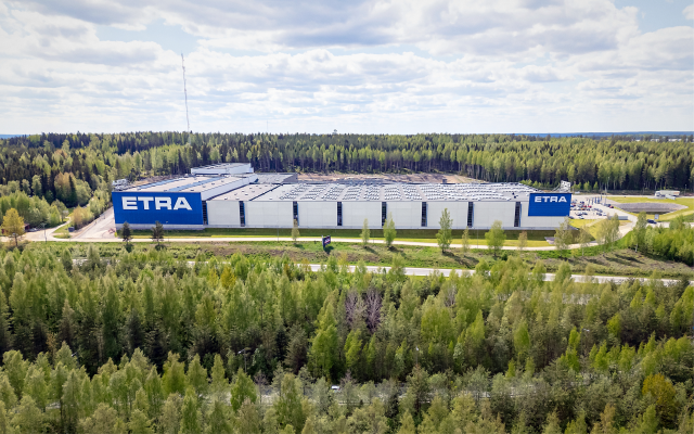 Ilmakuva Etran logistiikkakeskuksesta, jota ymp&auml;r&ouml;i laaja mets&auml;alue. Rakennuksen katolla on aurinkopaneelit ja sinivalkoiset ETRA-logot erottuvat selke&auml;sti.