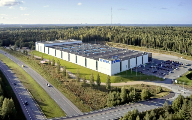 Exterior view of Etra's logistics center – a large, modern warehouse building with the company logo prominently displayed. In the background, an open sky and a spacious yard area.