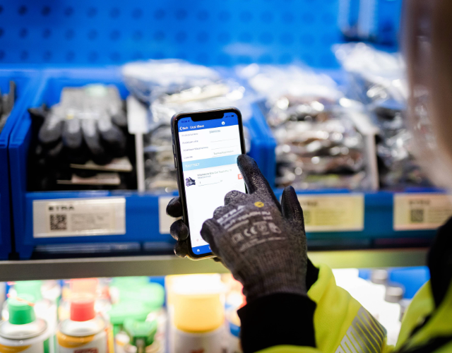 Työntekijä käyttää älypuhelinta hyllyn ääressä, jossa on Etran tuotteita ja varastokoodeja. Puhelimen näytöllä näkyy Etran mobiilisovellus