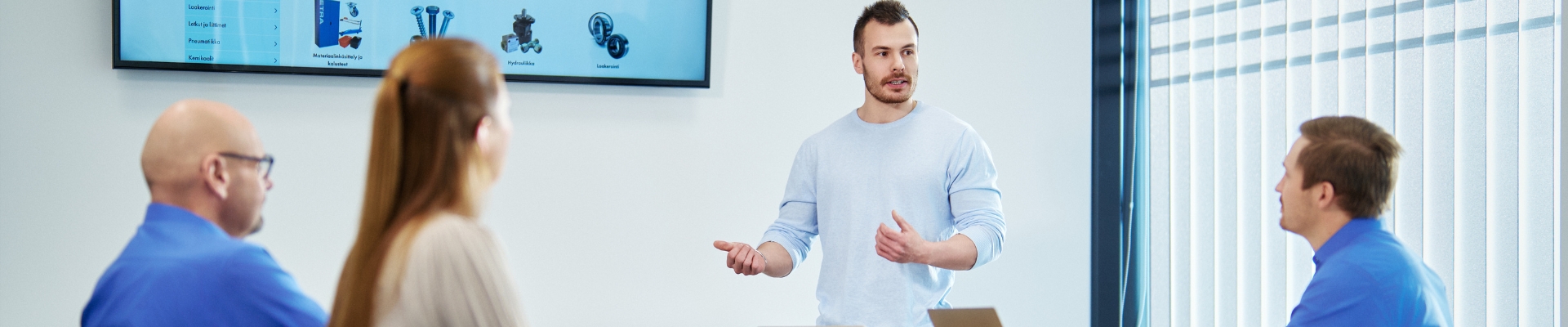 A presentation in a meeting room where a young man is standing and speaking to three seated individuals. In the background, a large screen displays a website showcasing various tools.