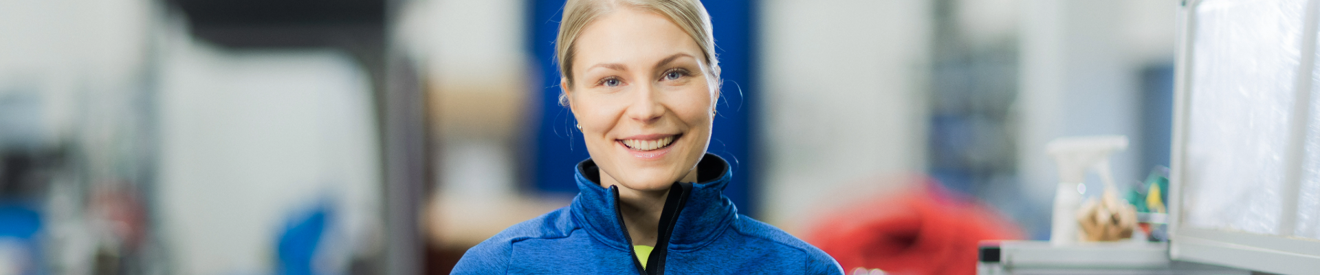 Hymyilevä nainen Etran työvaatteissa teollisessa ympäristössä. Kuvassa korostuu positiivinen ilmapiiri ja asiantuntevuus työympäristössä.