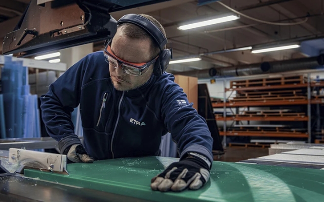 Etra employee machining a plastic sheet with a processing machine, wearing safety glasses, hearing protection, and gloves.