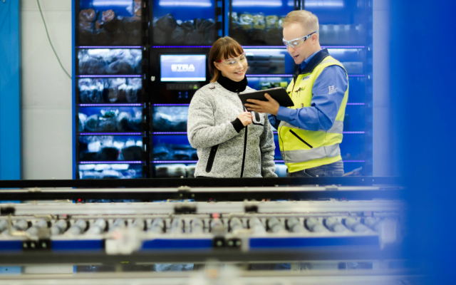 Kaksi henkilöä keskustelevat Etran palveluvaraston edessä, taustalla näkyy automaattisia varastokaappeja valaistuine hyllyineen. Etualalla kuljetinrullasto ja mies esittelee tablettia naiselle, molemmat käyttävät suojalaseja.