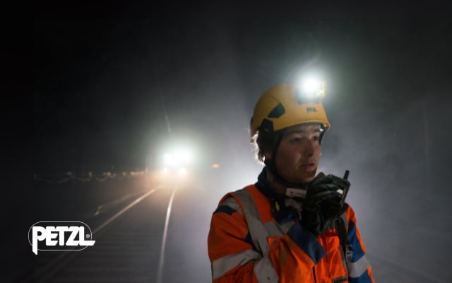 Työntekijä rautatietyömaalla pimeässä, pukeutuneena oranssiin huomiotakkiin ja keltaiseen kypärään, jossa on Petzl-otsalamppu. Hän puhuu radiopuhelimeen, ja taustalla näkyy valoja ja junarata.