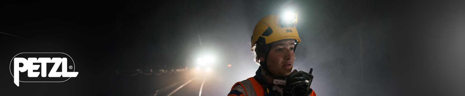 Työntekijä rautatietyömaalla pimeässä, pukeutuneena oranssiin huomiotakkiin ja keltaiseen kypärään, jossa on Petzl-otsalamppu. Hän puhuu radiopuhelimeen, ja taustalla näkyy valoja ja junarata.
