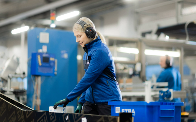 Nainen työskentelee tuotantolinjalla kuulonsuojaimet päässään ja käsineet kädessään, pakaten pieniä tuotteita ETRA-merkityistä laatikoista sinisiin muovilaatikoihin. Taustalla näkyy koneita ja teollista tuotantoympäristöä.