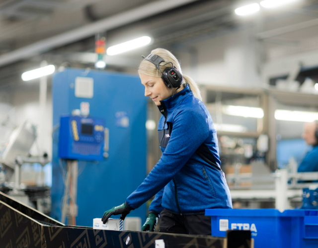 Etra-anställd som kontrollerar en produkt vid en förpackningsmaskin i en produktionsmiljö, iförd varselväst och keps.