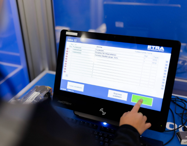 Henkilö käyttää ETRAN itsepalvelupäätettä, jonka näytöllä näkyy varastojärjestelmän raportointinäkymä. Kosketusnäytöllä valitaan tuotteita ja vahvistetaan tapahtuma "Valmis"‑painikkeella.