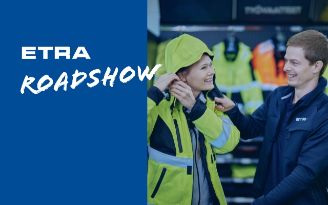 Kaksi hymyilevää henkilöä sovittamassa työturvallisuusvaatteita ETRA Roadshow -tapahtumassa, taustalla näkyy värikkäitä työvaatteita.