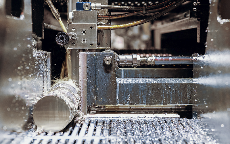 A metallic saw machine cuts a round metal bar in a production facility. The cutting process generates metal shavings, and cooling fluid flows around the blade. The pipe is secured in a sturdy holder inside the machine.