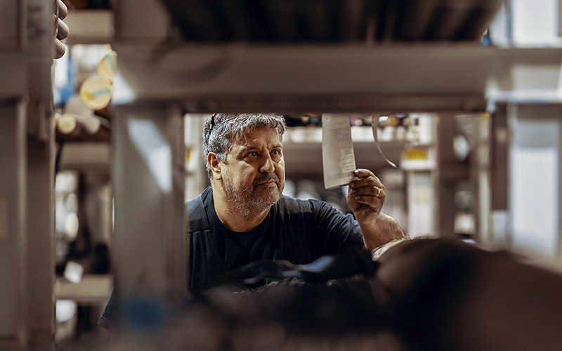 A worker in a metal storage area examines a label attached to a metal pipe between the shelves. The shelves contain metal bars and pipes of various sizes. In the background, rows of storage shelves are visible.