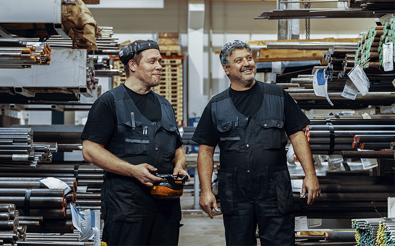 Two workers stand in a metal storage area between shelves filled with metal bars and pipes of varying lengths and thicknesses. One worker is holding a remote control, and both are wearing black work clothes and vests. In the background, a yellow crane is 
