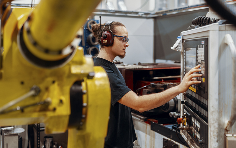 A worker operates the control panel of a CNC machine in an industrial facility. The person is wearing safety glasses and hearing protection. In the foreground, a yellow robotic arm is visible.