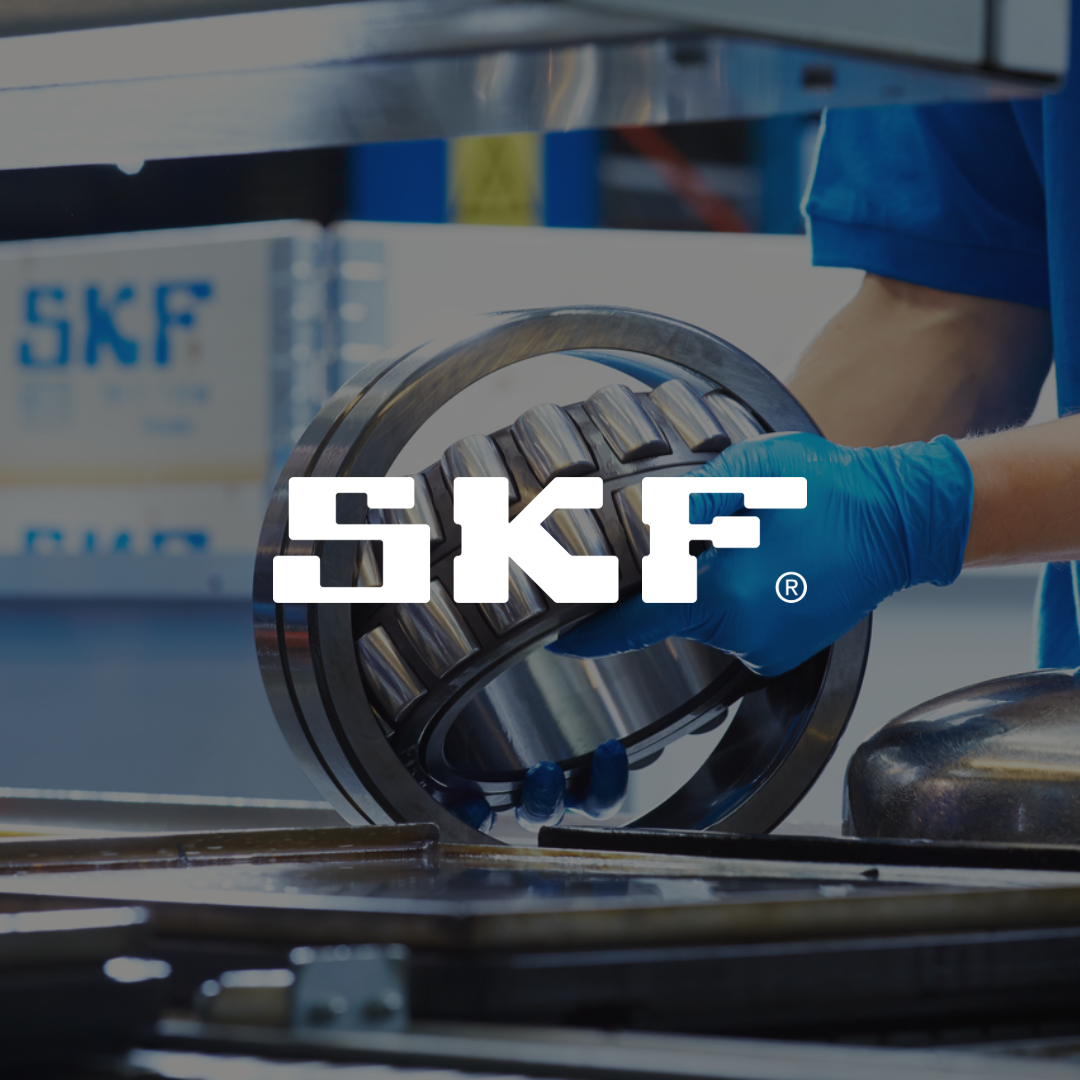 Henkilö sinisissä suojakäsineissä käsittelee suurta laakeria työpisteellä. Taustalla näkyy SKF-logoja laatikoissa. Kuvan keskellä on suuri valkoinen SKF-logo.