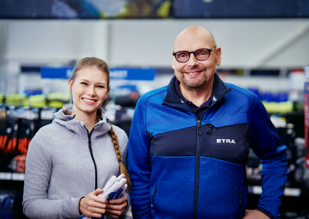 Kaksi henkilöä hymyilee kameralle myymäläympäristössä. Vasemmalla oleva nainen on pukeutunut harmaaseen huppariin ja pitelee työ- tai suojakäsineitä. Oikealla oleva mies käyttää sinistä ETRA-logolla varustettua takkia ja silmälaseja. Taustalla on hyllyjä.