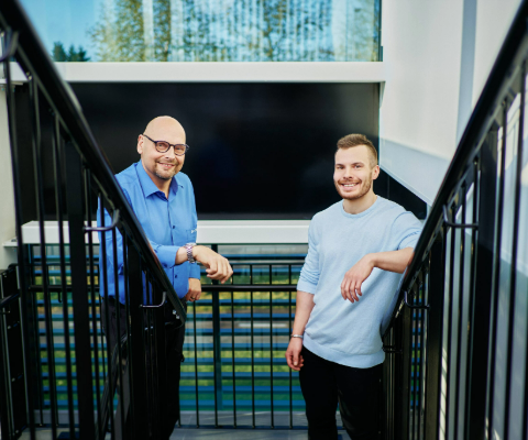 Two smiling men stand in a stairwell inside a modern office building.