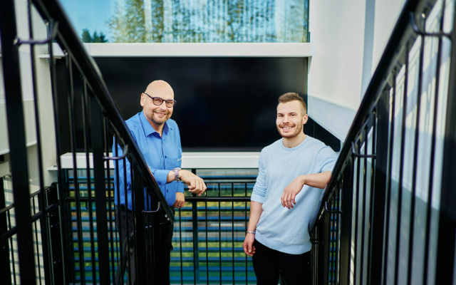 Two smiling men stand in a stairwell inside a modern office building.