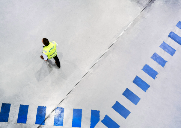 Henkilö kävelee harmaalla betonilattialla, jossa on sinisiä viivoja kaarevassa muodossa. Henkilöllä on yllään keltainen huomioliivi ja mustat housut. Kuva on otettu ylhäältä päin.