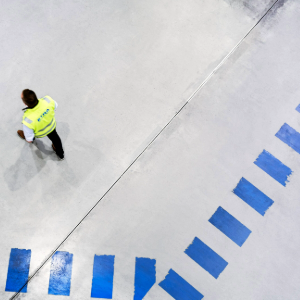 Henkilö kävelee harmaalla betonilattialla, jossa on sinisiä viivoja kaarevassa muodossa. Henkilöllä on yllään keltainen huomioliivi ja mustat housut. Kuva on otettu ylhäältä päin.