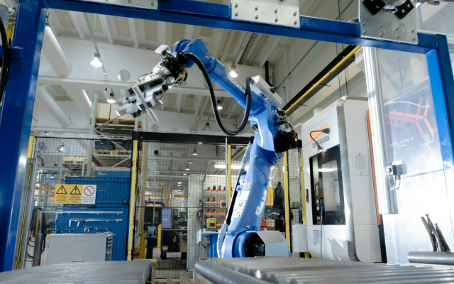 En industrirobot arbetar vid en produktionslinje i en modern fabriksmiljö, med maskiner och skyddsstängsel i bakgrunden.