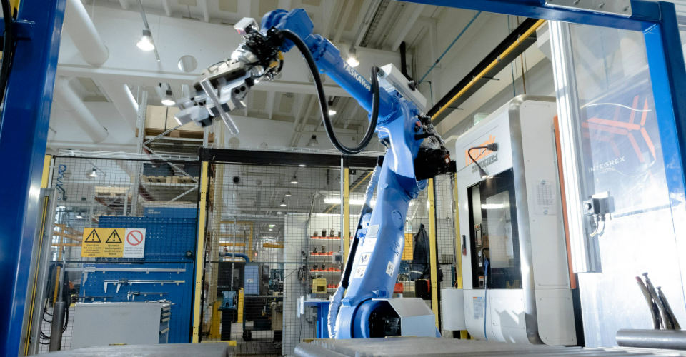 En industrirobot arbetar vid en produktionslinje i en modern fabriksmiljö, med maskiner och skyddsstängsel i bakgrunden.