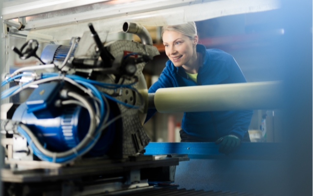 The image shows a person working in an industrial environment with various machines and equipment visible. There are also blue cables and metallic parts in the scene.