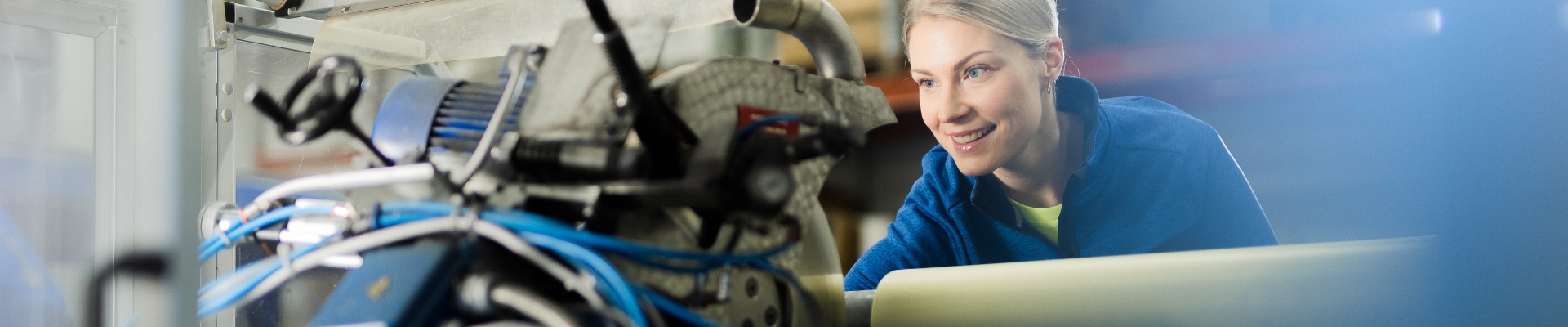 The image shows a person working in an industrial environment with various machines and equipment visible. There are also blue cables and metallic parts in the scene.