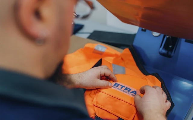 Worker finishing an orange workwear garment with the Etra logo using a heat press.