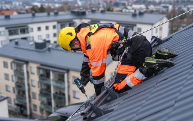 Kattoty&ouml;ntekij&auml; kaltevalla peltikatolla ja kiinnitt&auml;&auml; rakennetta porakoneella. Henkil&ouml;ll&auml; on p&auml;&auml;ll&auml;&auml;n huomiov&auml;rinen oranssi turvavaatetus, kyp&auml;r&auml; ja turvavaljaat, jotka on kiinnitetty k&ouml;ydell&auml; kattoon. Taustalla n&auml;kyy kerrostaloja.