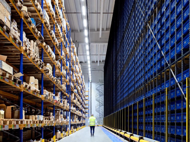 Logistiikkakeskuksen korkea varastohalli, jossa työntekijä heijastinliivissä kävelee suuren kuormalavahyllystön ja automaattivaraston välissä.