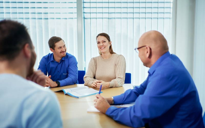 A group of people having a meeting in a bright office space. A woman and a man are smiling at each other, while two other participants are listening.