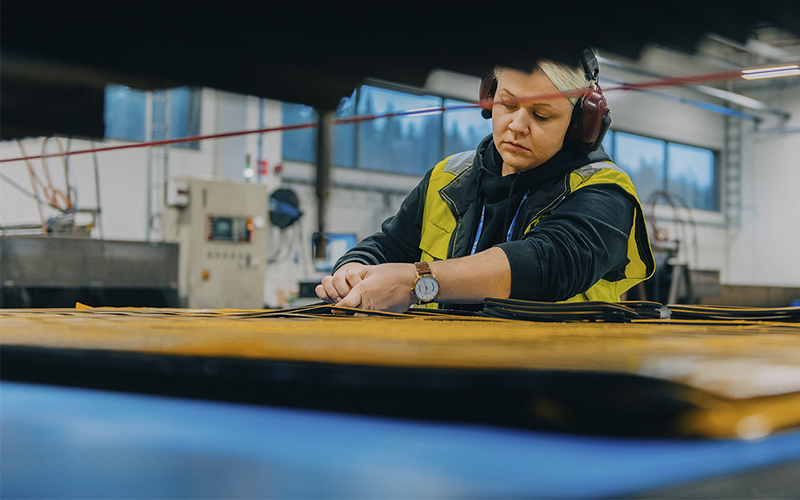 A person wearing a high-visibility vest and ear protection is working at a table in an industrial or workshop setting. Large windows and various equipment and machinery are visible in the background.