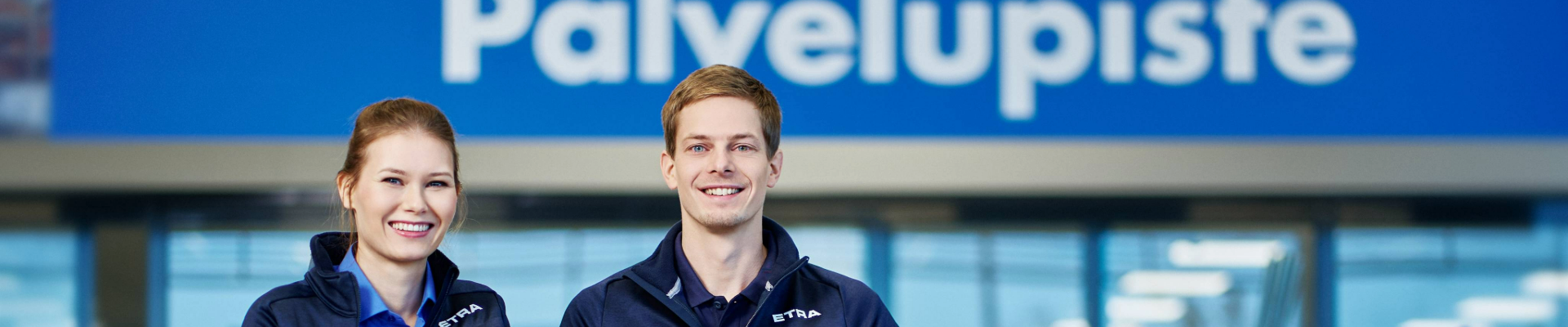 Två leende kundservicerepresentanter står framför en servicestation. De har på sig blå arbetskläder, och en modern butiksmiljö med ljus belysning syns i bakgrunden. 