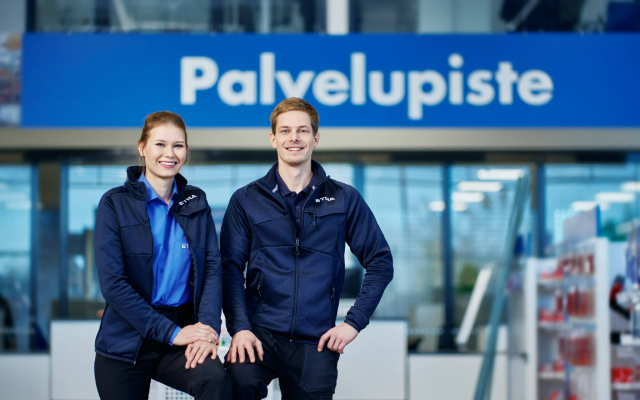 Two smiling customer service representatives stand in front of a service point. They are wearing blue work uniforms, and a modern store environment with bright lighting is visible in the background.